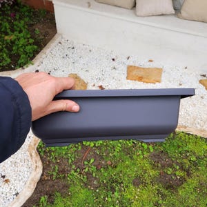 Op de afbeelding: Een rechthoekige, donkergrijze plantenbak, vastgehouden door een persoon. De plantenbak heeft een eenvoudig, modern ontwerp met rechte zijkanten en een iets verhoogde basis. De achtergrond omvat groen, witte kiezels en een witte muur.