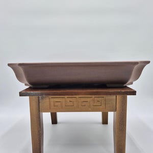 Puede incluir: Una maceta rectangular marrón para bonsái se encuentra sobre un pequeño soporte de madera. La maceta tiene lados suavemente curvados y una base plana. El soporte tiene cuatro patas y un patrón de greca tallado en el frente.