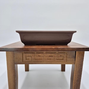 Op de afbeelding: Een bruine rechthoekige bonsai pot staat op een houten tafel. De tafel heeft een Grieks sleutelmotief en wordt ondersteund door vier vierkante poten. De pot is donkerbruin.