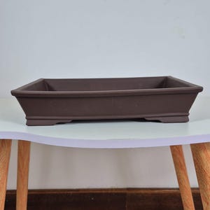 Puede incluir: Una maceta rectangular marrón para bonsái con un perfil bajo. La maceta está hecha de cerámica o un material similar y se encuentra sobre una mesa blanca con patas de madera. Sus dimensiones son de aproximadamente 30 cm de largo, 20 cm de ancho y 8 cm de profundidad.