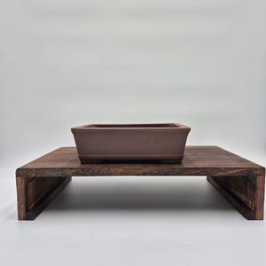 Könnte beinhalten: Ein rechteckiger, brauner Bonsai-Topf steht auf einem dunklen Holzständer. Der Ständer hat eine rechteckige Oberseite und eine untere Ablage, die von abgewinkelten Holzstützen getragen wird. Topf und Ständer stehen vor einem weißen Hintergrund.
