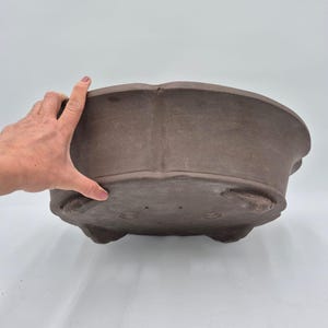Könnte beinhalten: Ein brauner, ovaler Bonsai-Topf mit gewelltem Rand und drei kleinen Füßen. Der unglasierte Keramiktopf hat eine matte Oberfläche und ist für das Pflanzen kleiner Bäume oder Sträucher konzipiert. Der Topf wird von einer Hand gehalten.