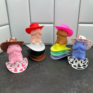 Puede incluir: Cuatro figuras novedosas, cada una con un sombrero de vaquero, descansan sobre bases apiladas y coloridas. Las figuras son en tonos rosa, beige y azul. Los sombreros son rojos, rosas, marrones y blancos con lunares rojos. Las bases son blancas, negras, marrones, amarillas y moradas.