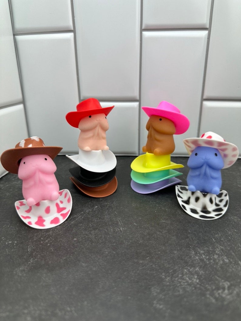 Puede incluir: Cuatro figuras novedosas, cada una con un sombrero de vaquero, descansan sobre bases apiladas y coloridas. Las figuras son en tonos rosa, beige y azul. Los sombreros son rojos, rosas, marrones y blancos con lunares rojos. Las bases son blancas, negras, marrones, amarillas y moradas.
