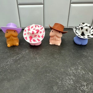 Puede incluir: Cuatro figuras novedosas, cada una con un sombrero de vaquero &uacute;nico. Los sombreros son morados, rosas con lunares blancos, marrones y con estampado de vaca en blanco y negro. Las figuras son de varios tonos de piel y azules.