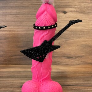 May include: A pink figurine with a white fuzzy hairpiece, a black studded belt, and a black glitter guitar. The figurine is shaped like a human male body.