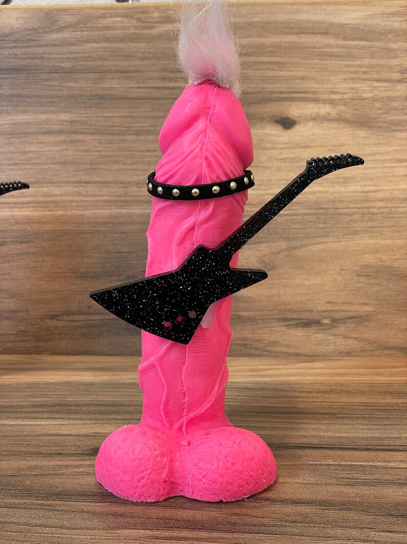 May include: A pink figurine with a white fuzzy hairpiece, a black studded belt, and a black glitter guitar. The figurine is shaped like a human male body.