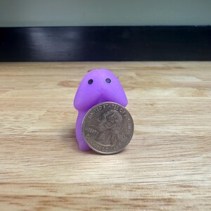 Puede incluir: Una peque&ntilde;a figura morada, de estilo caricatura, con dos ojos negros, de pie junto a una moneda de veinticinco centavos estadounidense. La figura tiene una cabeza redondeada y un cuerpo cil&iacute;ndrico. La moneda es de color plateado y presenta un perfil de George Washington.