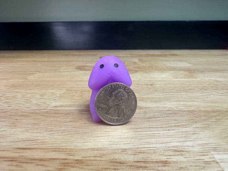 Puede incluir: Una peque&ntilde;a figura morada, de estilo caricatura, con dos ojos negros, de pie junto a una moneda de veinticinco centavos estadounidense. La figura tiene una cabeza redondeada y un cuerpo cil&iacute;ndrico. La moneda es de color plateado y presenta un perfil de George Washington.