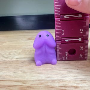 Puede incluir: Una peque&ntilde;a figura de color lavanda con un cuerpo redondeado y dos peque&ntilde;os ojos negros. La figura mide aproximadamente 2,5 cm de altura y se mide con una regla morada.