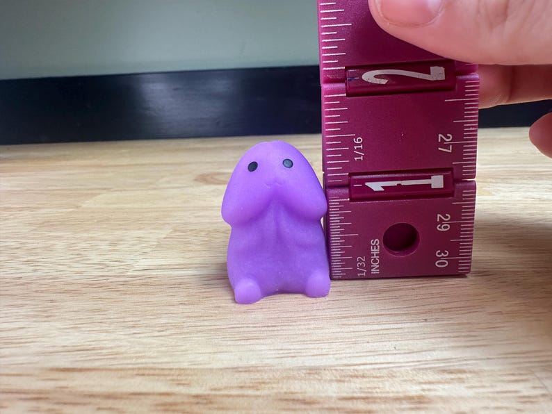 Puede incluir: Una peque&ntilde;a figura de color lavanda con un cuerpo redondeado y dos peque&ntilde;os ojos negros. La figura mide aproximadamente 2,5 cm de altura y se mide con una regla morada.