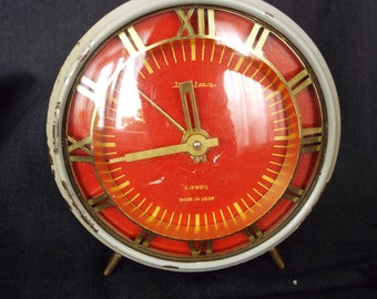 May include: A vintage red and white alarm clock with gold Roman numeral markings and a glass face. The clock has a 4 jewels inscription and says "Made in USSR".