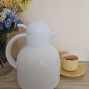 May include: A white thermal carafe with a rounded body, a curved handle, and a lever-operated lid. Beside the carafe sits a small, pale yellow ceramic teacup on a matching saucer. The design is simple, featuring a smooth, glossy finish throughout.