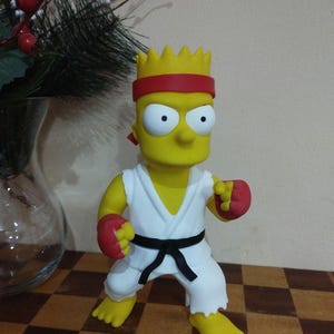 May include: A yellow Bart Simpson figurine in a karate outfit. He has a red headband, white karate gi, black belt, and red gloves. The figurine is posed in a fighting stance on a checkered surface.