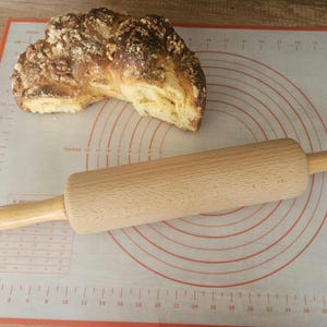 Puede incluir: Un rodillo de madera descansa sobre una esterilla de silicona para hornear con círculos concéntricos y marcas de medición. Una hogaza de pan con una corteza dorada y texturizada se encuentra en la parte superior. La esterilla tiene medidas en cm.