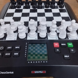 May include: A black and white electronic chess set with black and white chess pieces arranged on the board. The set has a built-in display and buttons for various functions. The brand name "ChessGenius" is visible.