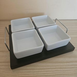 May include: A serving tray with four square white ceramic bowls. The bowls are set on a black rectangular tray with a silver metal handle. The tray is on a light wood surface.