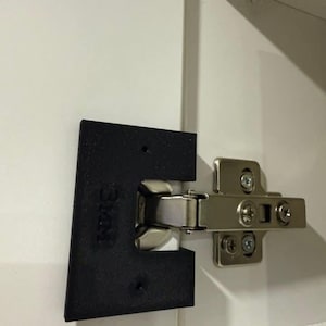 May include: A close-up of a cabinet hinge and a black plastic mounting plate. The hinge is silver and attached to a white cabinet door. The mounting plate has the text "S&M" printed on it.