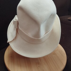 Peut inclure: Un chapeau cloche crème avec une couronne arrondie et un large bord. Un nœud en tissu assorti orne le côté. Le chapeau est présenté sur une surface en bois ronde.