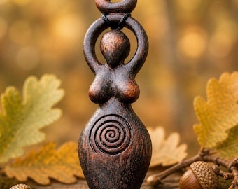 May include: A dark brown wooden pendant necklace featuring a stylized female figure with arms raised and a spiral design on the torso. The pendant is suspended from a black cord. The background includes acorns and autumn leaves.