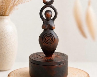 May include: A dark brown wooden statue of a female figure with arms raised, topped with a crescent moon. The statue sits atop a round, dark brown box with carved geometric designs. The box is on a wooden slice.