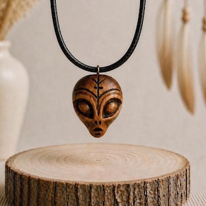 Peut inclure: Un collier avec pendentif en bois représentant un extraterrestre. Le pendentif marron est sculpté avec des traits détaillés et est suspendu à un cordon noir. Il est présenté sur une tranche de bois.