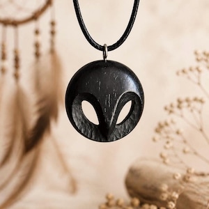 Può includere: Una collana con pendente nero con un design di gufo in legno intagliato. Il pendente rotondo ha due ritagli a forma di occhio e un becco. La collana ha un cordino nero e una chiusura color argento. Lo sfondo è beige neutro.
