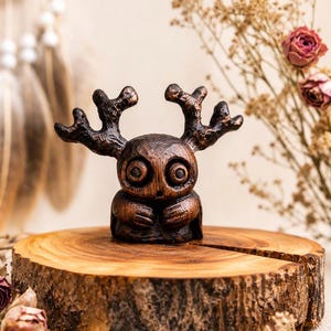 Peut inclure: Une petite figurine sculptée brun foncé présentant une tête ronde avec de grands yeux circulaires et des bois texturés proéminents sur le dessus. La figurine a un corps compact avec les mains jointes devant, arborant une finition boisée.