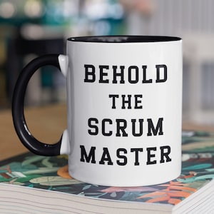 May include: White coffee mug with a black handle and black rim. The mug has the text "Behold the Scrum Master" printed in black.