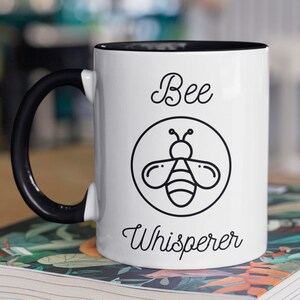 May include: White ceramic mug with a black handle and black rim. The mug has a black and white illustration of a bee with the text "Bee Whisperer" printed on it.