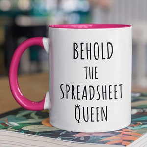 Puede incluir: Taza de cerámica blanca con borde y asa de color rosa. La taza tiene un texto negro que dice "Behold the Spreadsheet Queen".