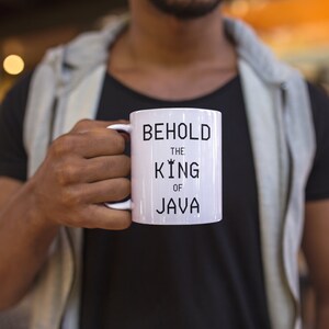 以下が含まれることがあります： 黒い文字で「Behold the King of Java」と書かれた白いセラミックマグカップです。