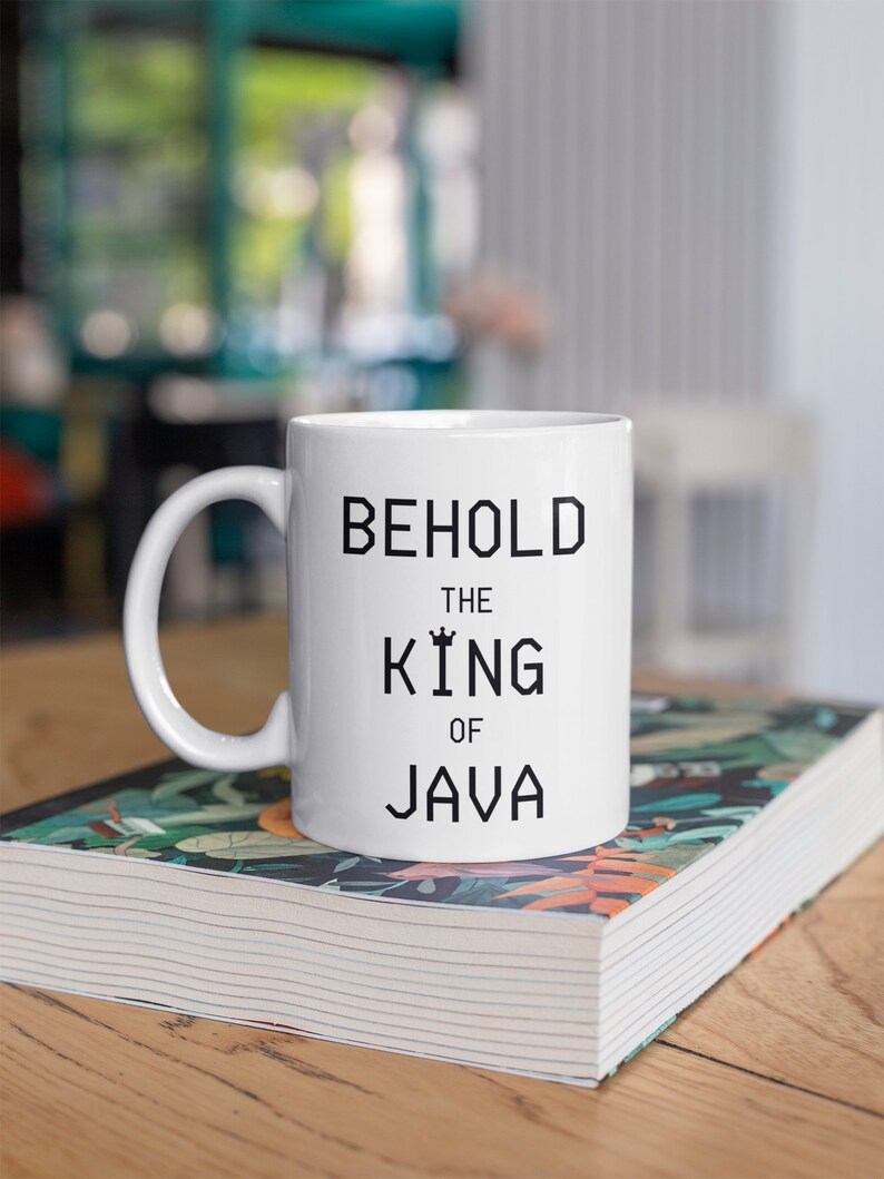 以下が含まれることがあります： 黒い文字で「Behold the King of Java」と書かれた白いセラミックマグカップです。