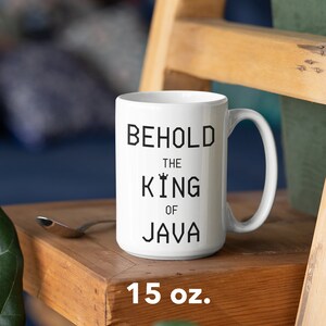 以下が含まれることがあります： 白いセラミック製のマグカップに、黒い文字で「Behold the King of Java」と書いてあります。マグカップは15オンスで、木製の表面の上に置かれています。