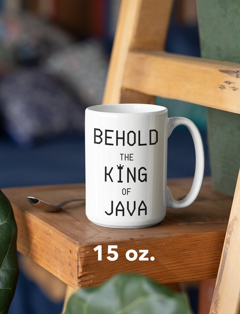 以下が含まれることがあります： 白いセラミック製のマグカップに、黒い文字で「Behold the King of Java」と書いてあります。マグカップは15オンスで、木製の表面の上に置かれています。