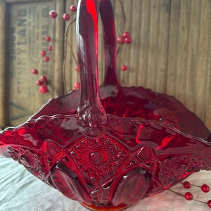 May include: A red glass basket with a scalloped edge and a diamond pattern. The basket has a single handle and is made of clear glass with a red tint.