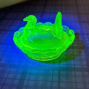 May include: A neon green glass dish in the shape of a duck sitting in a nest. The dish has a scalloped edge and a duck head and tail. The glass glows brightly against a dark background.