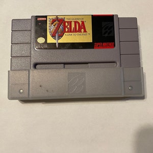 May include: A gray Super Nintendo cartridge for the game "The Legend of Zelda: A Link to the Past." The label features the game's title in red and gold, with a sword and shield graphic. The cartridge is rectangular.