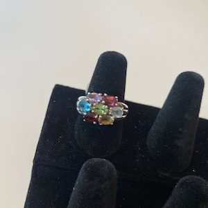 May include: A silver ring with a cluster of oval-shaped gemstones in various colors, including red, green, blue, and purple.