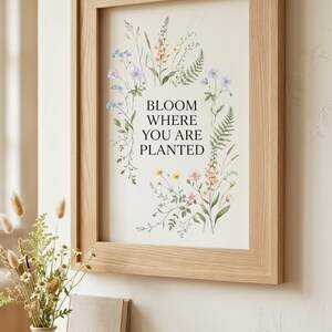 May include: A light wood framed wall art print featuring the black text BLOOM WHERE YOU ARE PLANTED centered on a cream background, surrounded by a delicate watercolor illustration of various wildflowers, ferns, and small green leaves.
