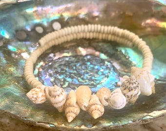 May include: A white shell bead bracelet with a row of small seashells attached. The bracelet is displayed on a iridescent abalone shell.