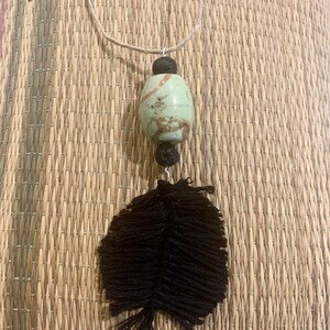 May include: A green and brown stone pendant with black lava beads hangs from a brown cord necklace. A black feather tassel dangles from the bottom of the pendant.