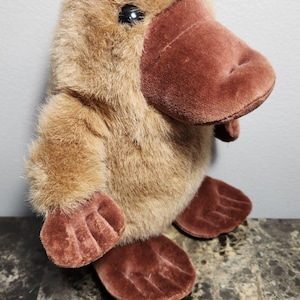 May include: A plush platypus toy featuring soft, light brown fur, a dark brown velvet-like bill, and matching dark brown webbed feet. The stuffed animal has a rounded body, a prominent flat bill, and a single visible black plastic eye.