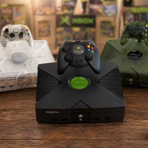 以下が含まれることがあります： 3台のXboxビデオゲームコンソールと対応するコントローラー。1台は黒で緑のロゴ入り、1台は透明、もう1台はHaloブランドの半透明の緑色です。各コンソールには前面ディスクドライブとコントローラーポートが備わっています。