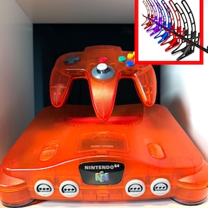 Könnte beinhalten: Orangefarbene, durchscheinende Nintendo 64-Konsole mit passendem Controller. Die Konsole hat das Nintendo 64-Logo auf der Vorderseite. Eine Auswahl an Controller-Ständern in verschiedenen Farben befindet sich in der oberen rechten Ecke.