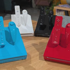 May include: Four Nintendo Wii game consoles in blue, white, black, and red, each paired with a matching Wii Remote and Nunchuk controller. The consoles are positioned upright with the controllers docked on top, displaying the Wii logo on each unit.