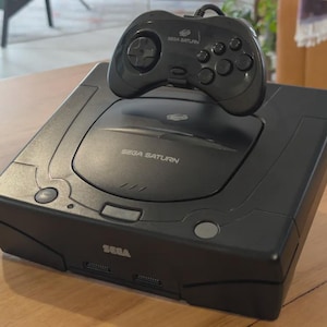 May include: A black Sega Saturn video game console with a matching black wired controller resting on top. The console features the Sega Saturn logo on the disc lid and the Sega brand name on the front panel, along with two controller ports.