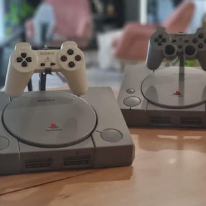 Peut inclure: Deux consoles de jeux Sony PlayStation grises avec des couvercles de disque circulaires et des ports de manette en façade. Chaque console dispose d'une manette assortie montée sur un support au-dessus du couvercle, affichant les logos Sony et PlayStation.