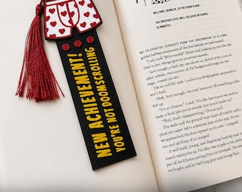 May include: A black rectangular bookmark with a red tassel, featuring a graphic of white boxer shorts with red hearts at the top. The text NEW ACHIEVEMENT! YOU'RE NOT DOOMSCROLLING is printed in yellow, with three small red circular icons above the text.