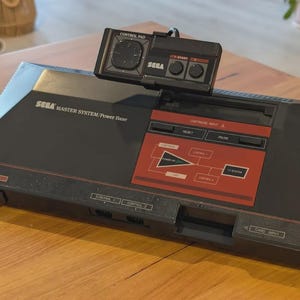 May include: A black Sega Master System Power Base video game console with a red control panel section, featuring a black rectangular control pad resting on top. The console includes labeled ports for cartridge input, card input, and two controllers.